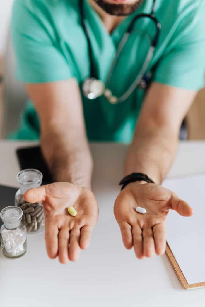 Unrecognizable male doctor wearing medical uniform showing pills to camera while working in modern clinic sitting at table with clipboard and stethoscope